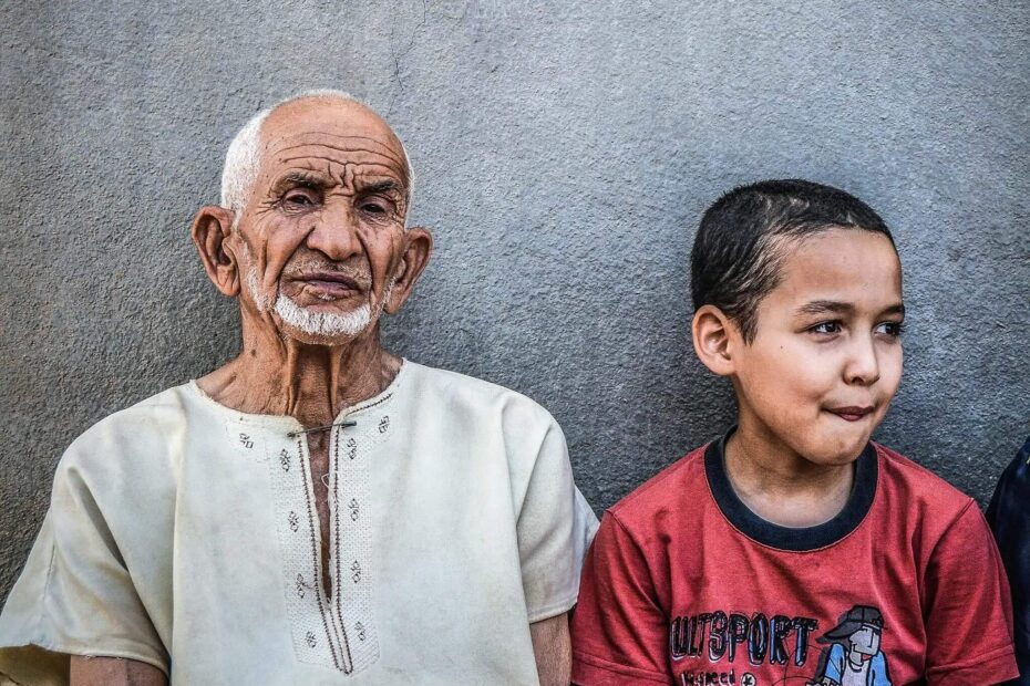 A young boy sitting next to his elderly grandfrather in front of a grey wall