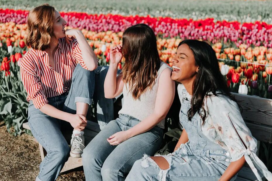 3 feminine presenting people talking and laughing while sitting on a bench in a field of flowers