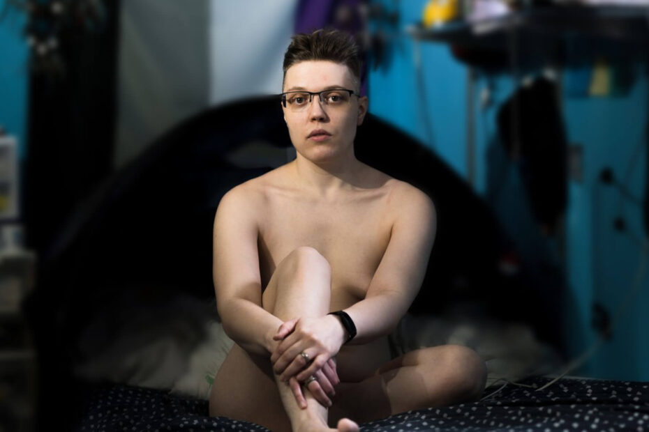 Arlo, a trans man sitting on the bed. He is naked though tastefully obscuring genitals and nipples via his pose using his arms and legs.