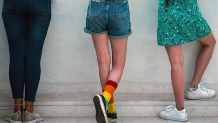 Three teens standing facing a wall. From right to left, there's a teenanger with dark skin wearing blue denim capri trousers, a white shirt, and white sneakers. The teenager in the middle is white and wearing a white shirt, blue denim shorts, black sneakers, and rainbow socks pulled up to mid-calf. The third teen has brown skin with long brown hair and is wearing a green floral dress with white sneakers.