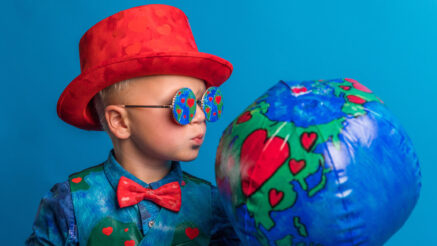 Young child in a red hat wearing a blue suit with red hearts, a red bow tie, and matching glasses, puckering up to an inflated ball that also matches the suit.